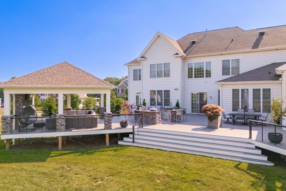 Spacious white house with a large deck, outdoor seating, and a covered gazebo. The deck features steps leading to a well-manicured lawn. The sky is clear and blue, enhancing the inviting atmosphere.