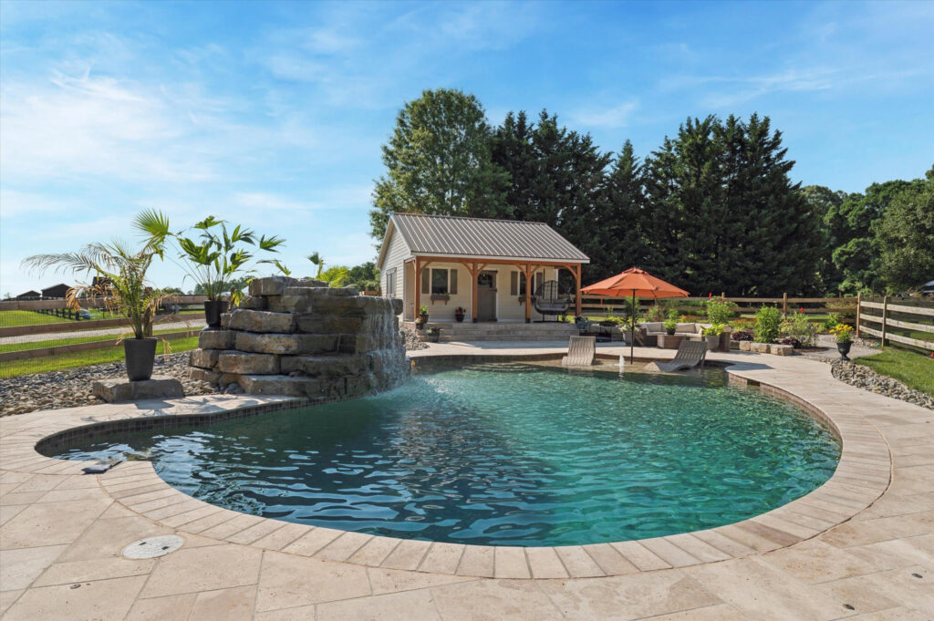 A backyard scene featuring a kidney-shaped swimming pool with a stone waterfall. Lounge chairs and an orange umbrella sit beside the pool. In the background, theres a small light-colored building and tall trees under a clear blue sky.