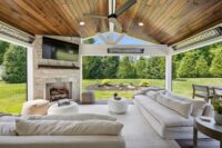 CO2DBB~1 | First State Building & Design A modern outdoor living space with white sofas, a stone fireplace, and a TV mounted above it. The room is open to a lush, green backyard with a well-manicured lawn and trees. The ceiling is wooden with a large fan. A dining table is on the right.