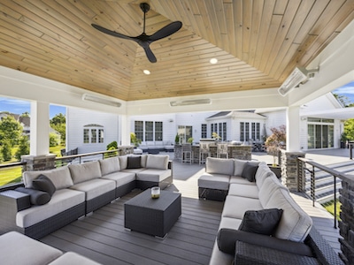 A spacious outdoor deck features a large, L-shaped gray sectional sofa with cushions around a dark coffee table. The area is covered with a wooden ceiling and ceiling fan. The background shows a white house with large windows and greenery.