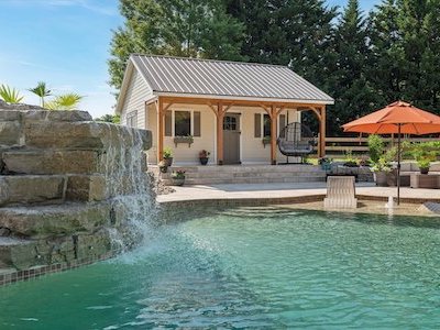 A backyard scene showing a small pool house with a metal roof and wooden columns. In front, theres a pool with a rock waterfall feature. An orange umbrella provides shade over a seating area. Trees and a clear sky are in the background.