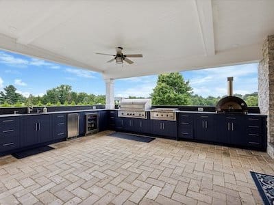 Outdoor kitchen with navy blue cabinets under a white ceiling. Features include a grill, oven, and refrigerator. A ceiling fan is above, and the area has brick flooring. Lush greenery is visible in the background.