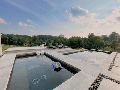 Modern outdoor pool with floating pool gadgets and inflatable rings, surrounded by stone tiles. Four dark lounge chairs on a wooden deck face the pool. Scenic, lush green landscape and partly cloudy sky in the background.