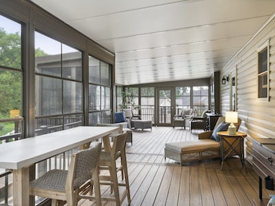 A bright, enclosed sunroom with large windows and wood flooring. It features a white table with woven chairs, a cushioned bench, a pair of armchairs, and a small side table with a lamp. The view outside shows greenery.