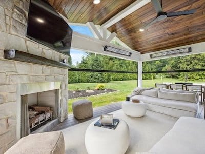 Luxurious outdoor living space with a stone fireplace, wall-mounted TV, and modern furniture. Features a wooden ceiling with a fan and large windows offering a view of the lush green landscape.