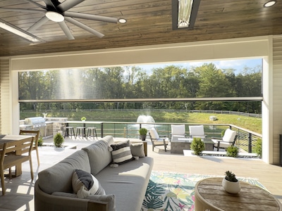 A spacious outdoor living area with a ceiling fan, featuring a sofa, dining table, and chairs. It overlooks a patio with sun loungers, a fountain, and a pond, surrounded by trees. Sunlight streams through the open space, creating a serene atmosphere.