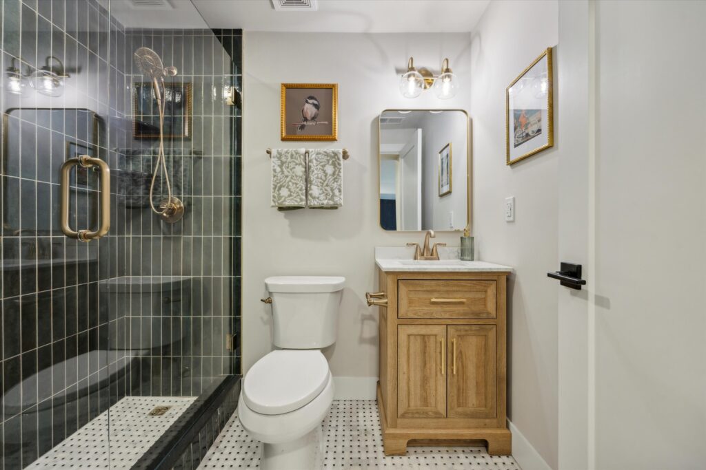 A modern bathroom with a glass shower enclosure and gold fixtures. It features a wooden vanity with a marble countertop, a rectangular mirror, and two wall lights. Decorative paintings and patterned black-and-white floor tiles complete the look.