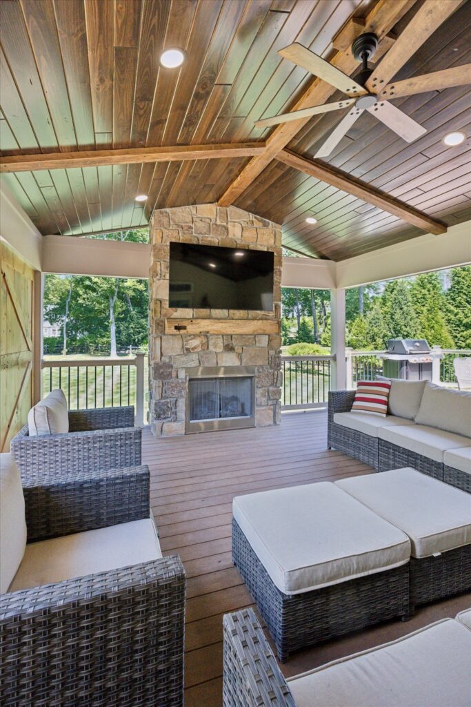 Bucks county A covered outdoor patio features wicker furniture with beige cushions, a stone fireplace with a mounted TV above it, a wooden ceiling with beams and a fan, and views of a green lawn and trees outside.