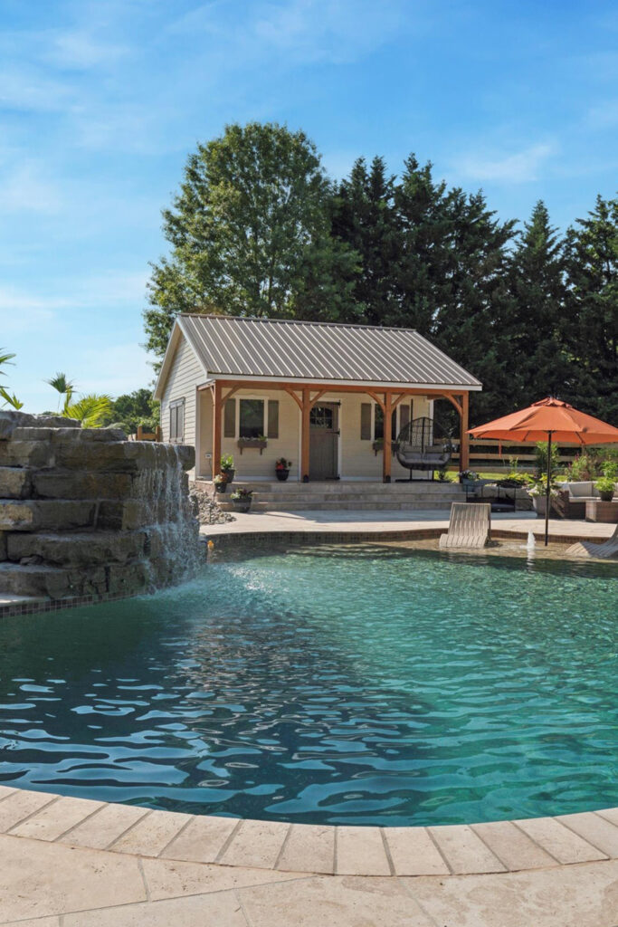 Cecil county A backyard scene with a swimming pool, waterfall feature, and a stone deck. In the background, there is a small covered patio with outdoor seating and potted plants, and an orange umbrella. Trees are behind the house.