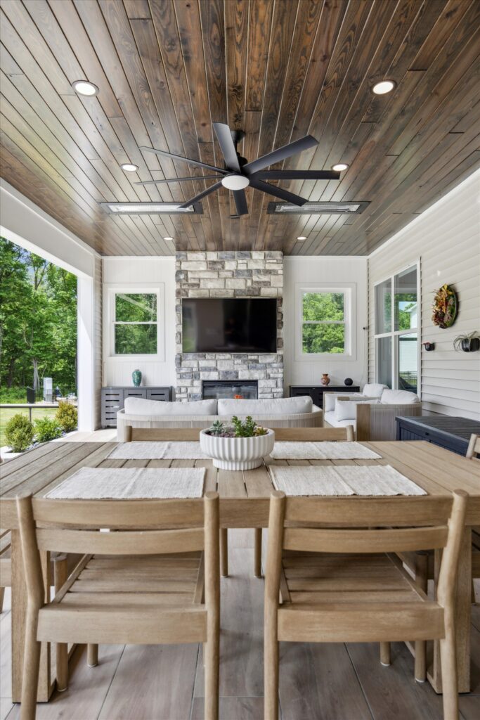 Chester county A modern covered patio with a wooden ceiling, large black ceiling fan, stone fireplace with a mounted TV, light wood dining table and chairs, white sofa, and view of a green yard through open windows.