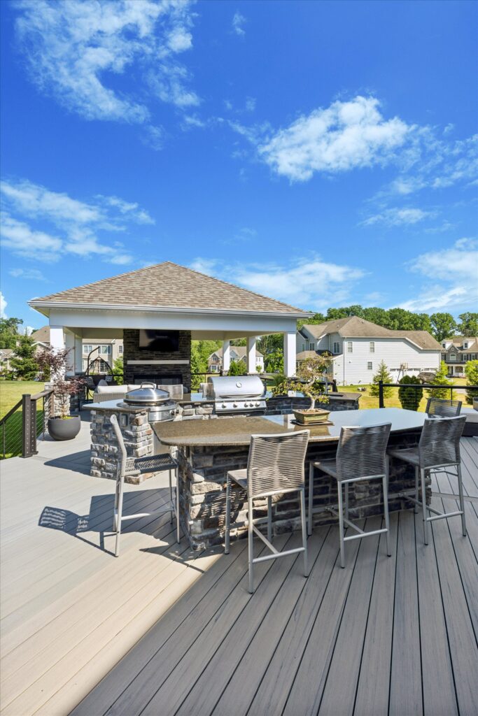 Delaware county A spacious outdoor deck with a built-in stone grill and bar area, metal barstools, and a covered patio. The scene is bright with blue skies and suburban homes visible in the background.