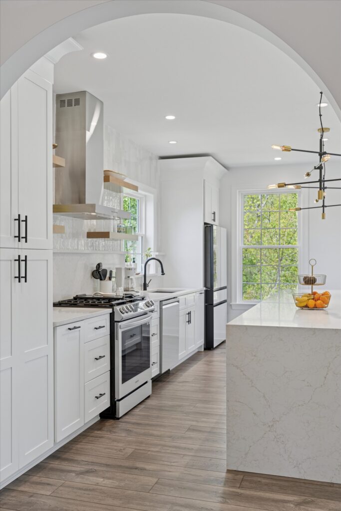 Montgomery county Modern white kitchen with stainless steel appliances, marble countertops, open shelves, large window with greenery outside, wooden floor, and a stylish ceiling light fixture.