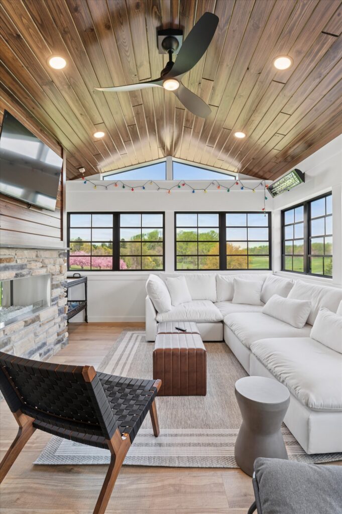 New Castle county A bright, modern sunroom with large windows, a white sectional sofa, a woven black chair, a wooden ceiling with recessed lights, a ceiling fan, and a wall-mounted TV above a stone fireplace.