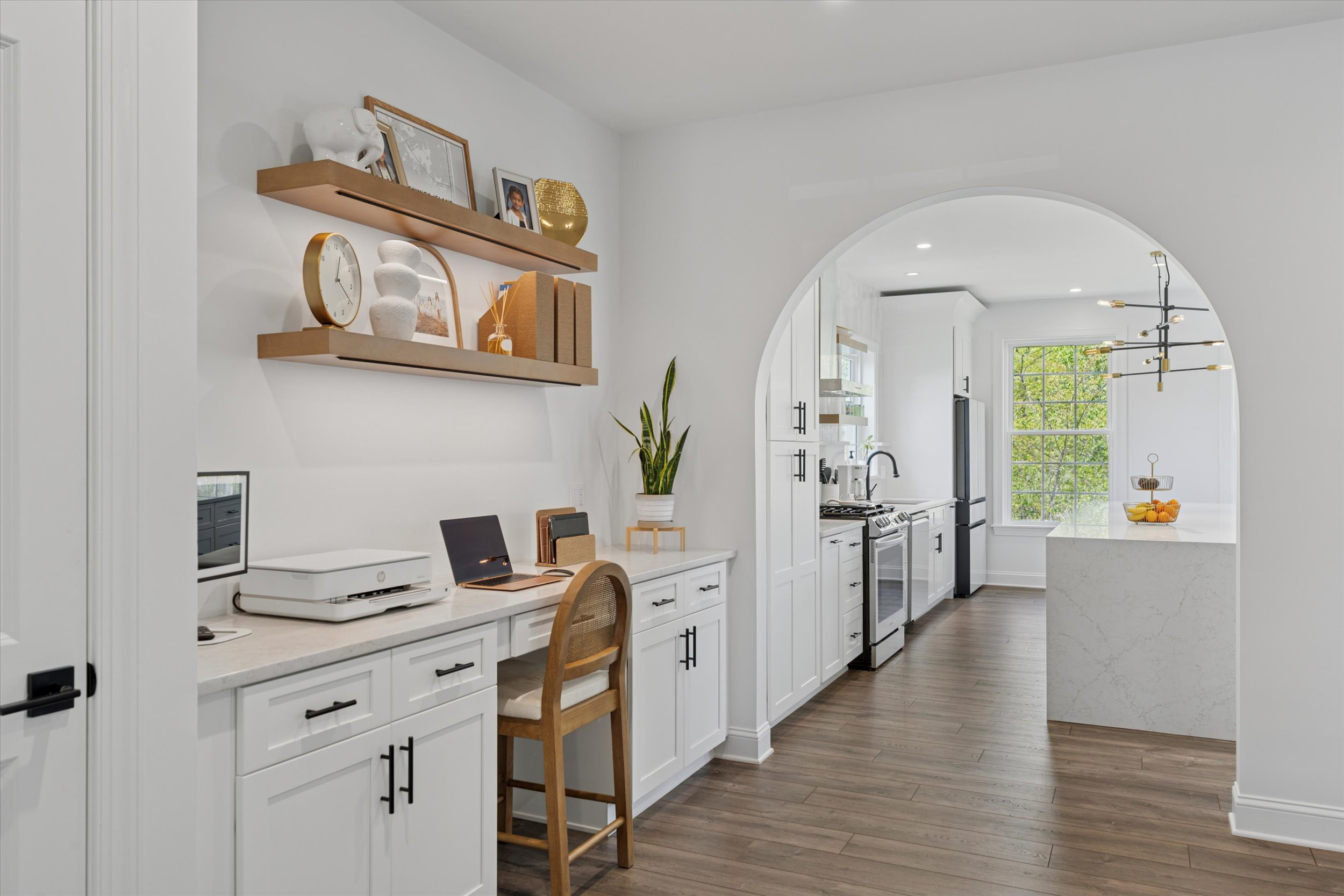 A modern home office nook with a wooden chair, white desk, shelves with décor, and a printer, opens to a bright kitchen with white cabinets, marble island, and a fruit bowl under a contemporary chandelier.