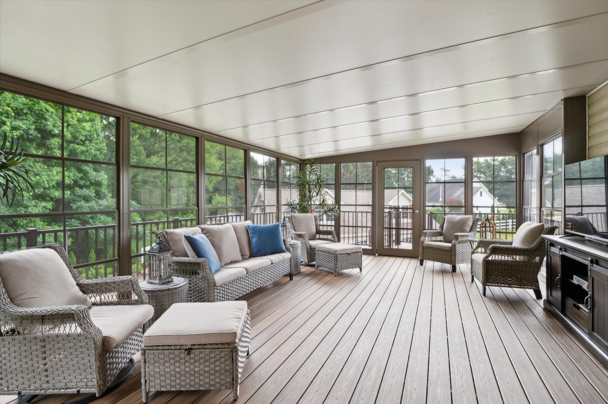 A spacious sunroom with wicker furniture, neutral cushions, and blue accent pillows. Large windows surround the room, offering a view of trees and houses outside. Wood-look flooring and plenty of natural light create an inviting atmosphere.