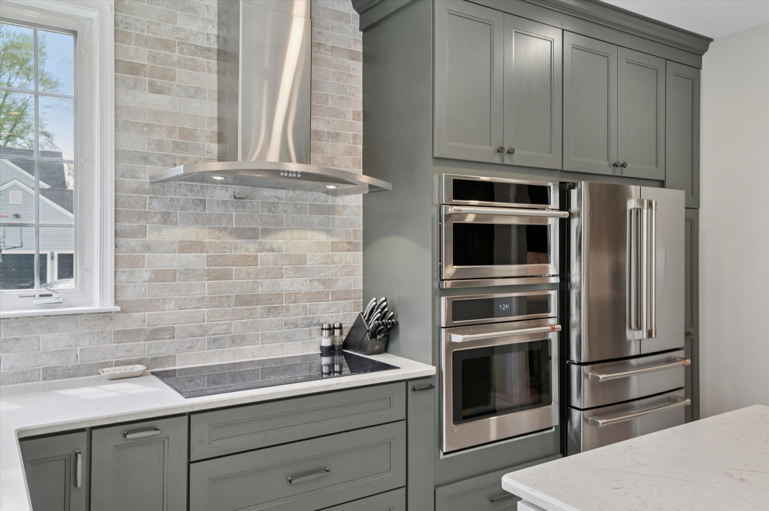Modern kitchen with green-gray cabinets, stainless steel appliances including a refrigerator, double wall oven, and range hood, white countertops, and a light brick backsplash. Knives and spices are on the counter.