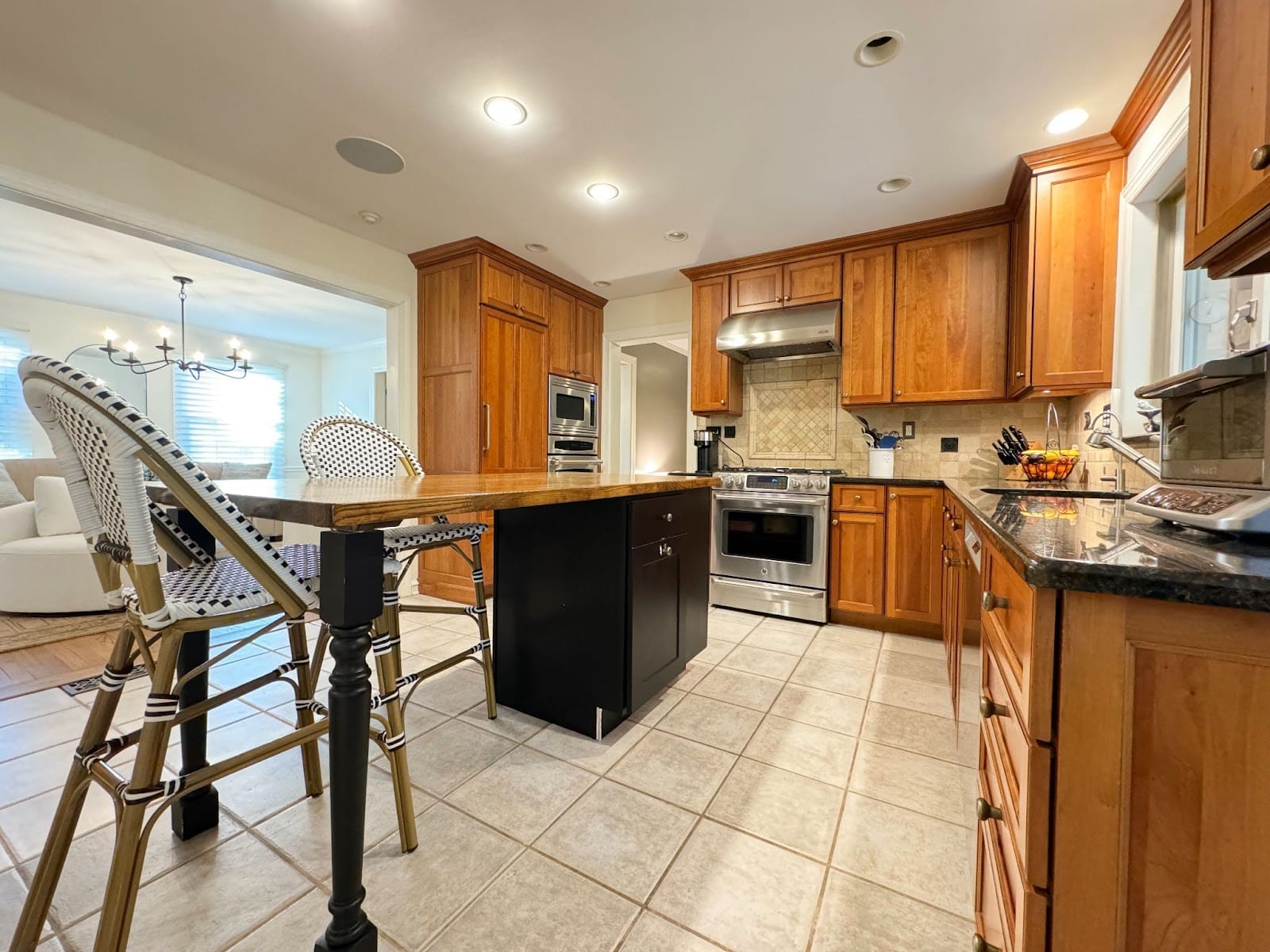 Spacious kitchen with wooden cabinets, a stainless steel stove, dark granite countertops, a central island with seating, and tile flooring. An adjoining dining area with a chandelier is visible in the background.
