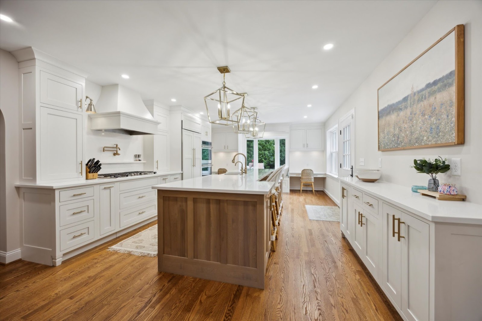Bright, modern kitchen with white cabinets, a wooden island, brass pendant lights, hardwood floors, and large windows bringing in natural light. A landscape painting hangs above the counter on the right wall.