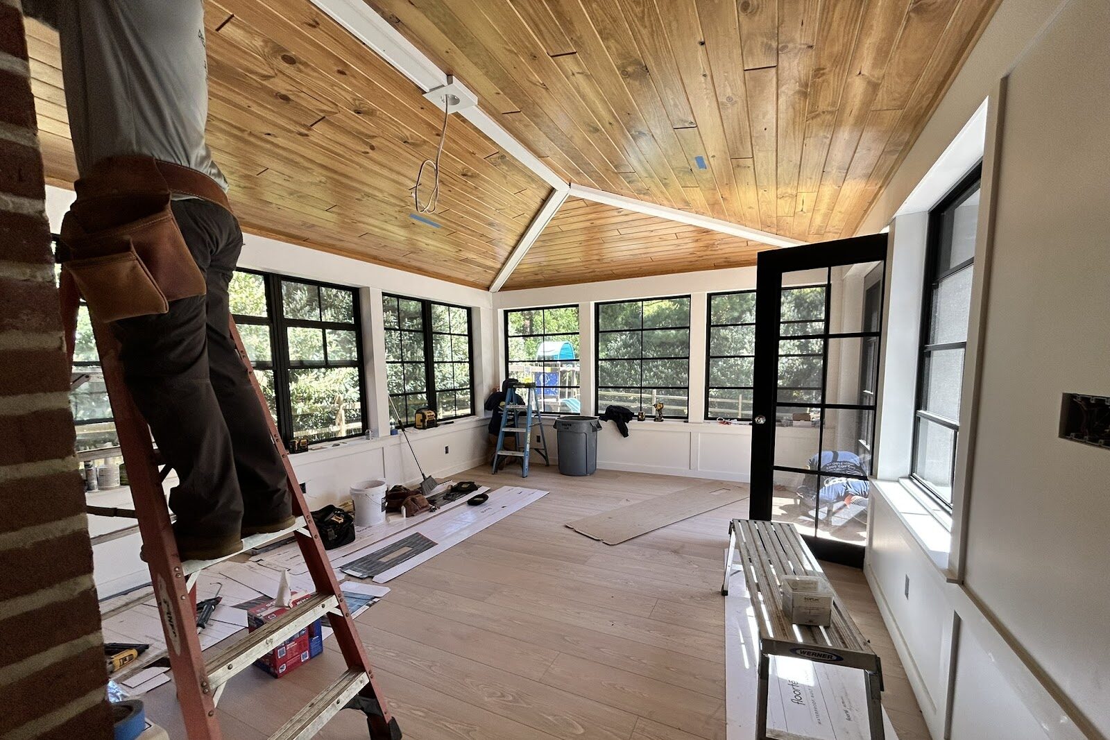 A person stands on a red ladder working on a ceiling in a bright, unfinished room with large windows, a wooden ceiling, and tools and materials scattered on the floor.