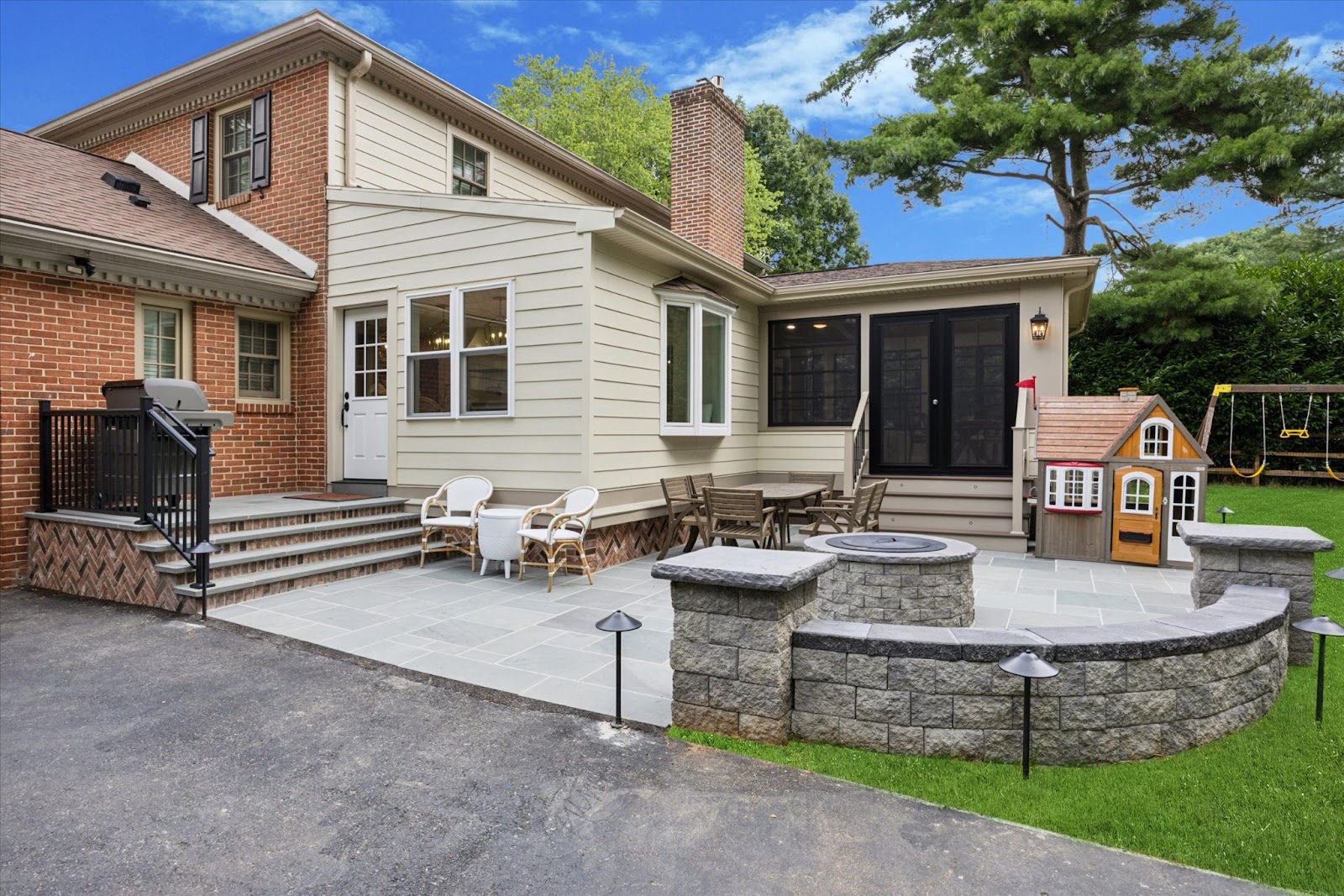 A spacious backyard patio features a fire pit with stone seating, outdoor furniture, a small playhouse, and a swing set. The home exterior combines brick and cream siding, with trees and a lawn in the background.
