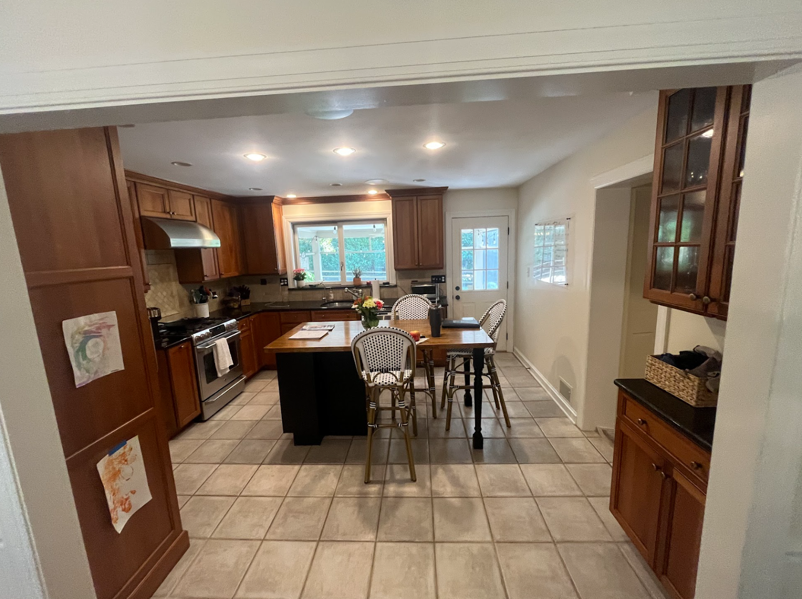 Spacious kitchen with wooden cabinets, a center island, and a dining table with four chairs. Large windows provide natural light. Drawings are displayed on the left wall, and kitchen appliances are visible on the countertops.