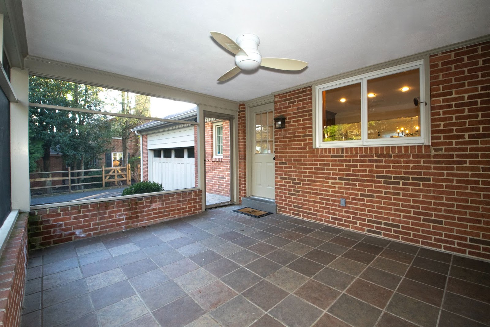 Enclosed porch with brick walls, tiled floor, ceiling fan, and large windows. Door leads to driveway and view of a garage and greenery outside. Warm indoor lighting visible through a window.