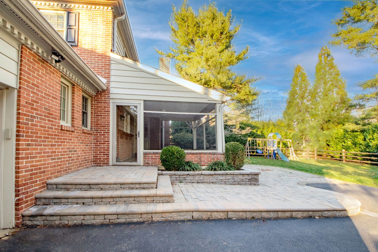 A brick house with a screened-in porch, stone steps, and a paved patio. There is green grass, trees, and a childrens playset with a slide in the fenced backyard under a blue sky.