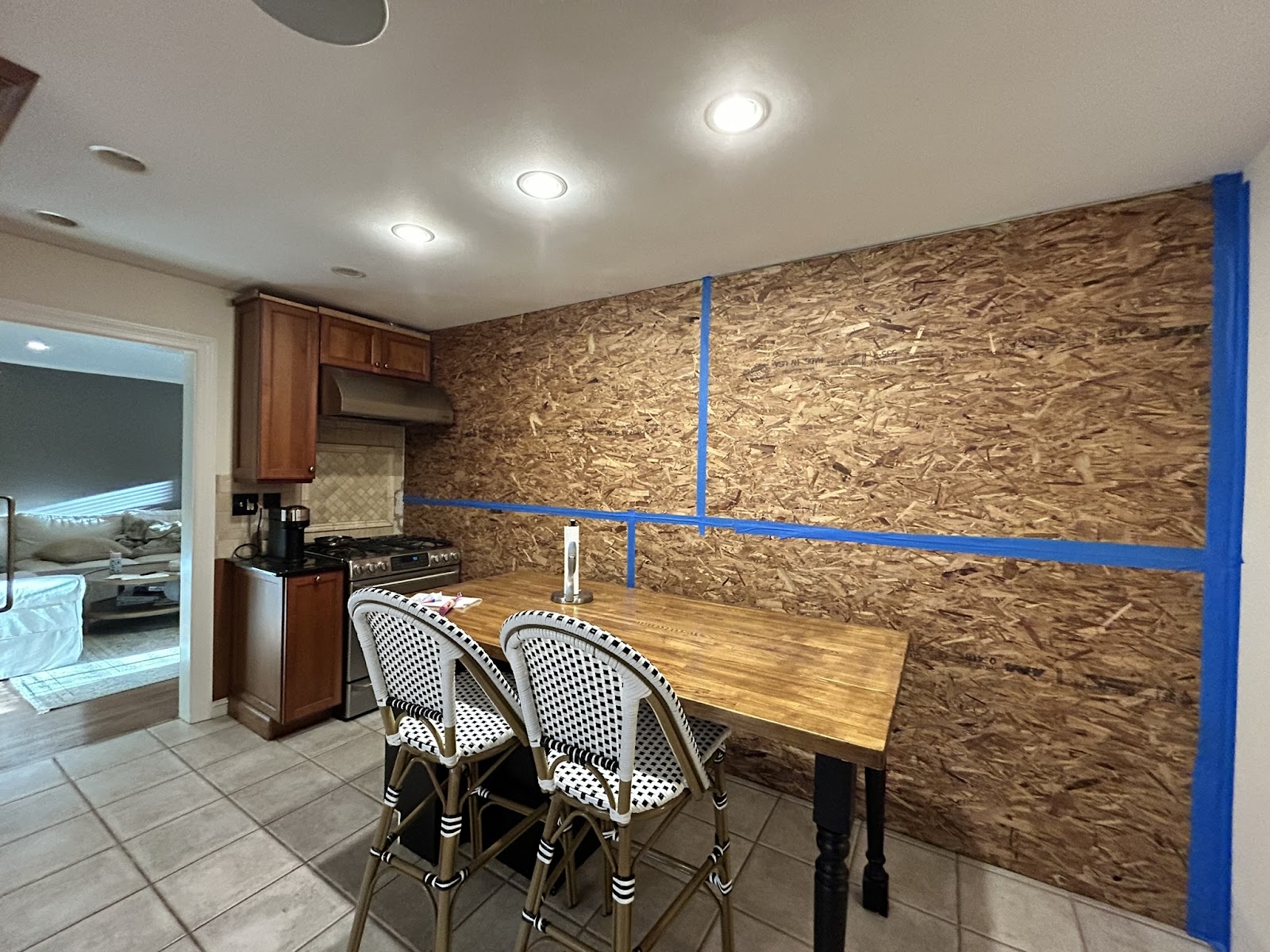 A kitchen with a wooden table, two chairs, a stove, and a coffee maker. The back wall is covered with plywood sheets and blue painter’s tape, suggesting ongoing renovation. A living room is visible through a doorway.