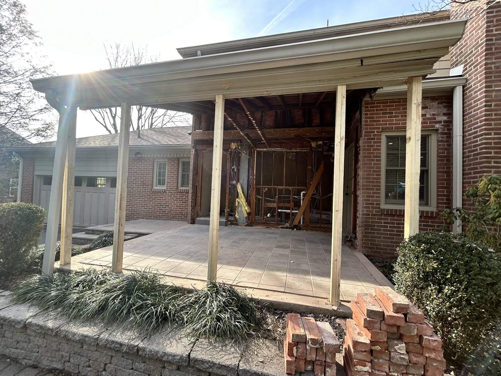 A partially demolished porch attached to a brick house, with exposed wooden beams and construction materials. A pile of bricks sits on the right, and the roof is supported by temporary wooden posts.