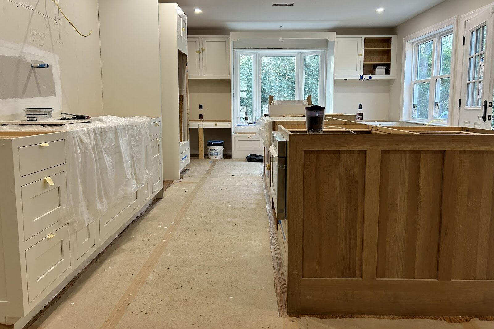 A modern kitchen under renovation with unfinished wooden and white cabinets, tools, and protective plastic covering surfaces. Large windows let in natural light, and the floor is bare, showing construction in progress.