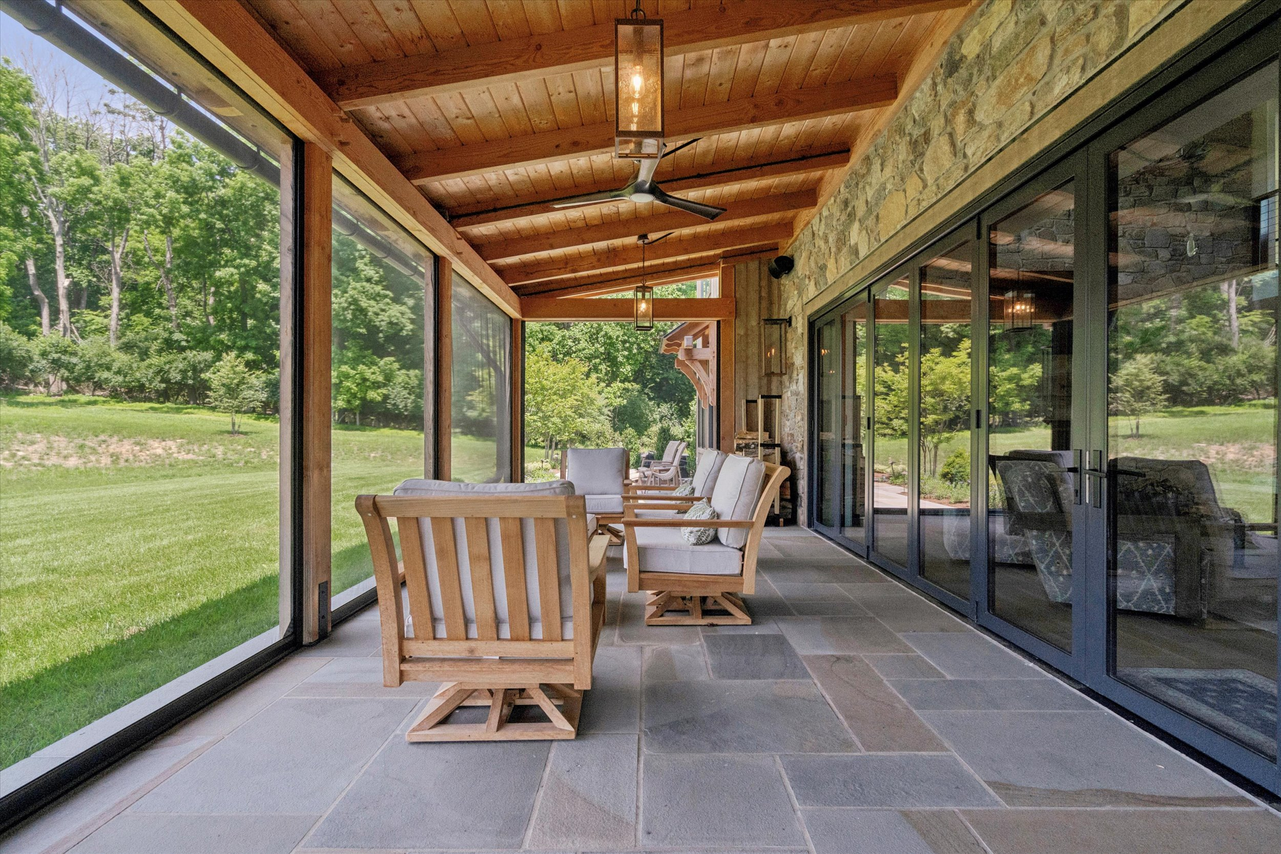 A covered patio with wooden ceiling beams, stone tile floor, and glass doors. Wooden chairs with cushions face a view of green grass and trees, creating a relaxing outdoor space.