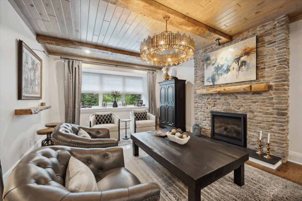 A cozy living room with tufted leather sofas, a large wooden coffee table, a stone fireplace, chandelier, artwork above the mantel, and a window with gray curtains and two chairs overlooking greenery outside.