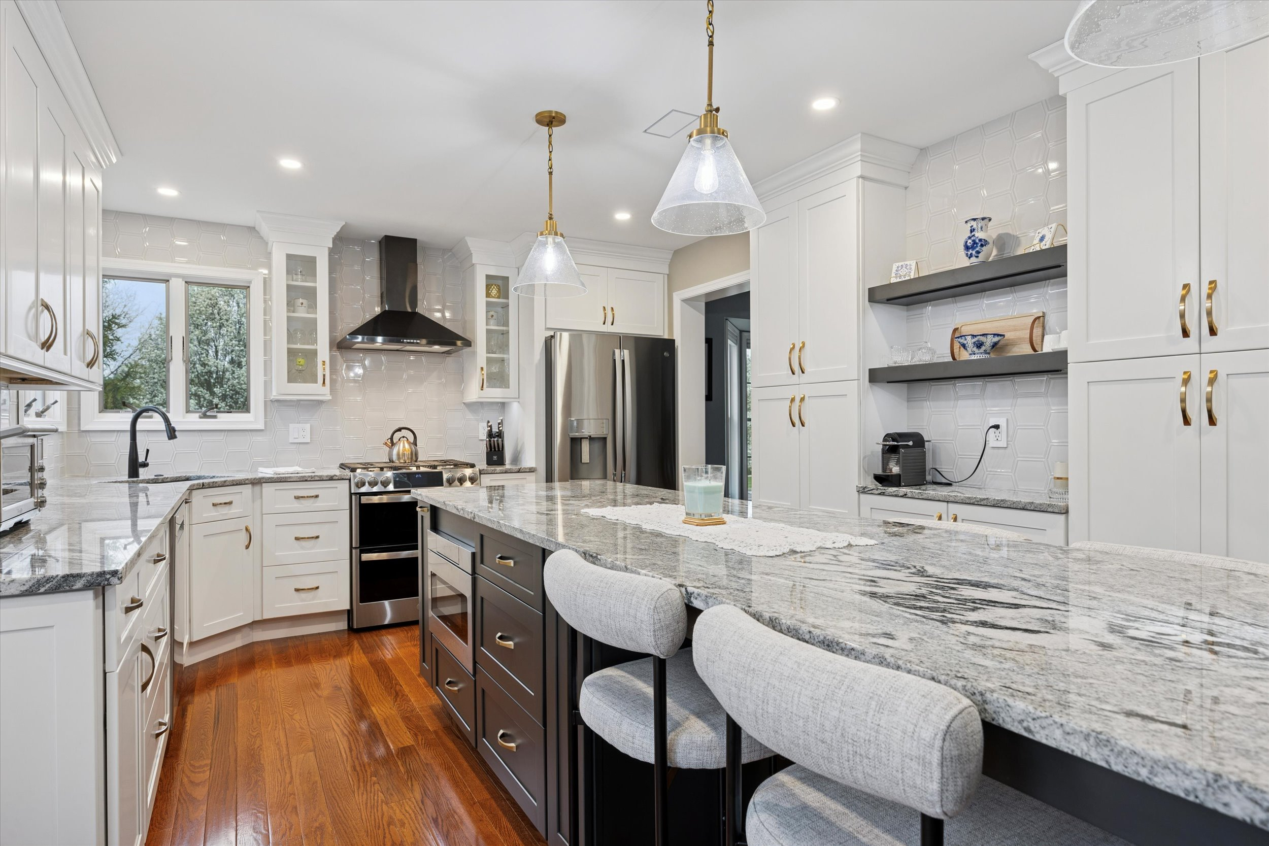 Modern kitchen with white cabinets, stainless steel appliances, a large gray granite island with barstools, wooden floors, pendant lights, open shelves, and a black range hood beneath a window.