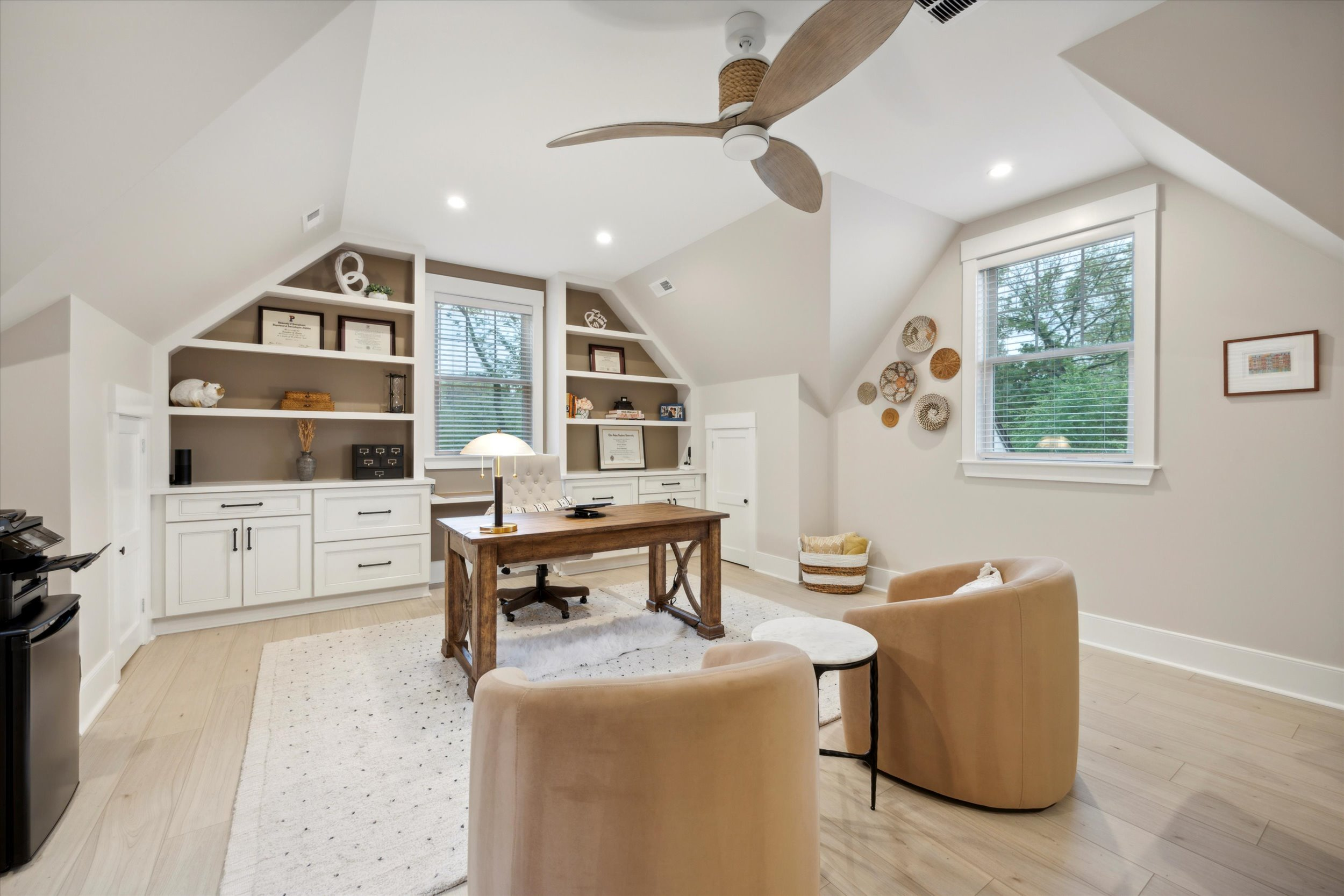 A cozy home office with sloped ceilings features a wooden desk, two beige armchairs, built-in white shelves, decorative baskets on the wall, a ceiling fan, and a window letting in natural light.