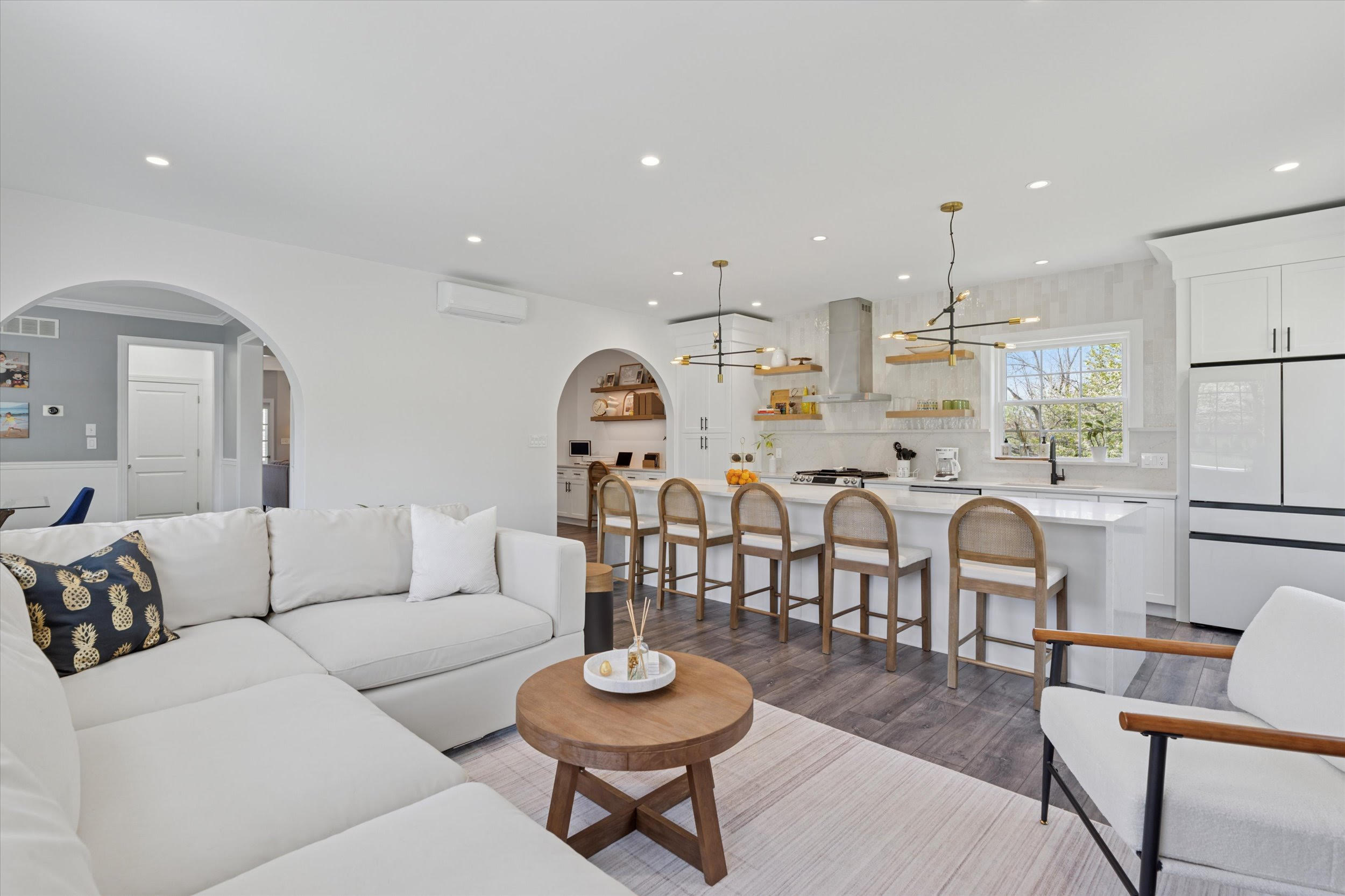 Modern open-concept living room and kitchen with white walls, neutral furniture, a large sectional sofa, wooden coffee table, four barstools at a kitchen island, pendant lights, and arched doorways.