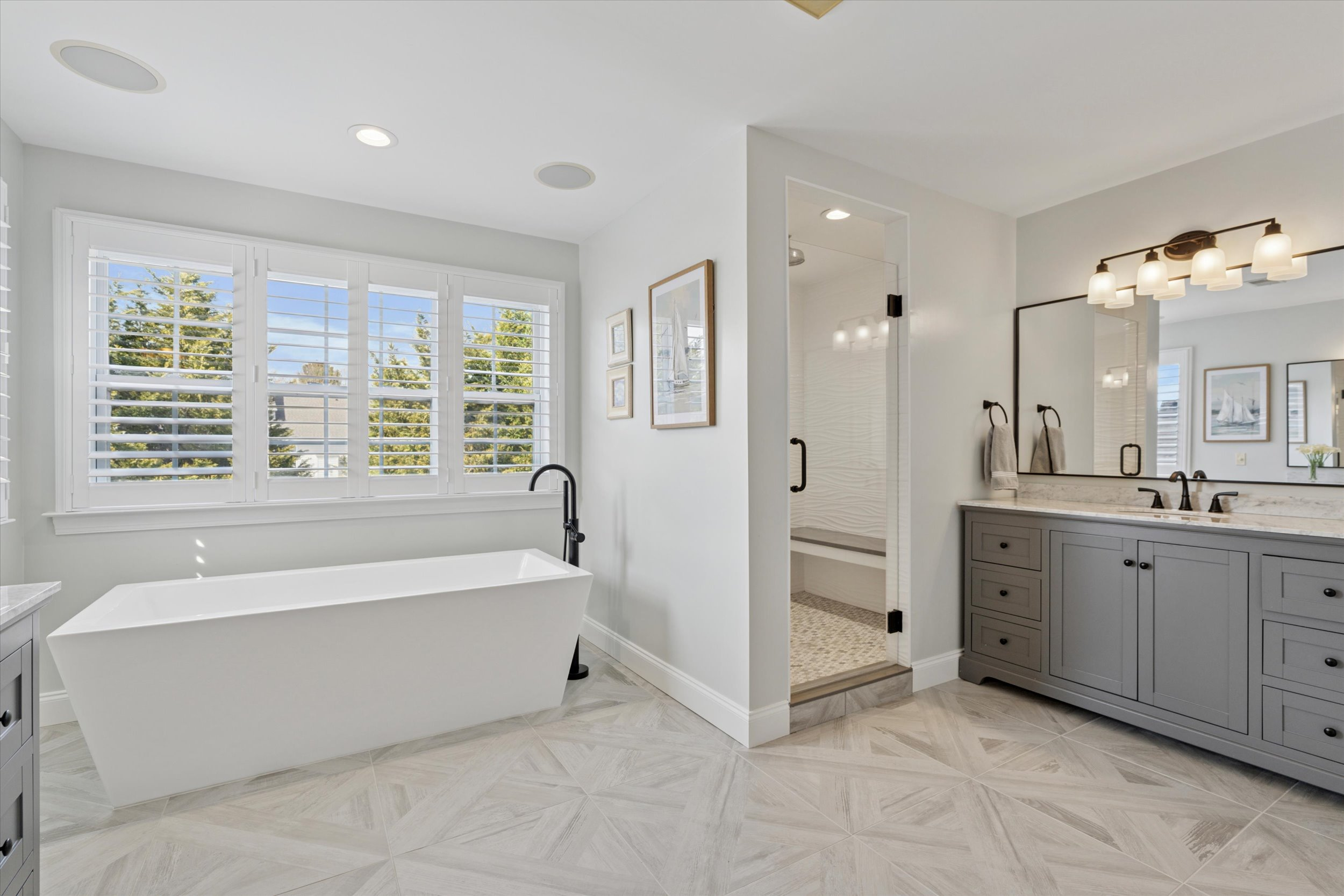 Bright, modern bathroom with a white freestanding tub near shuttered windows, double-sink gray vanity with marble top, a large mirror, and a glass-enclosed walk-in shower with a bench. Light tile flooring throughout.