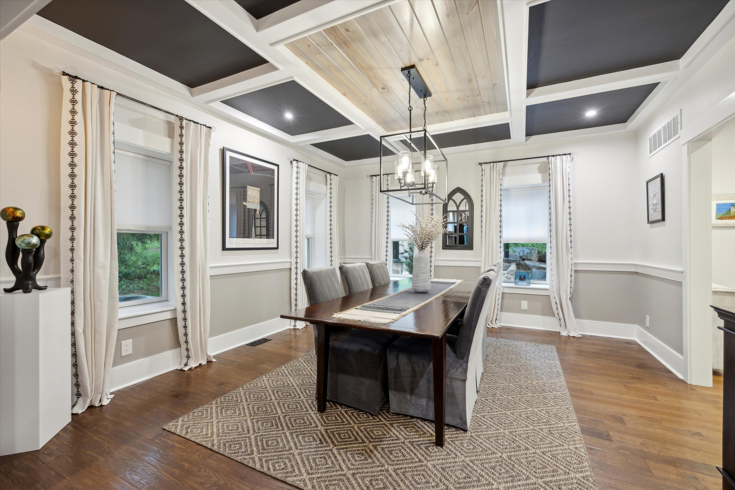 Elegant dining room with a long wooden table, gray upholstered chairs, patterned rug, coffered ceiling with wood accents, modern chandelier, large windows with white curtains, and decorative wall art.