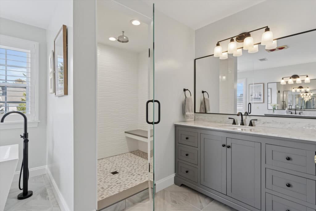 Modern bathroom with a glass-enclosed shower featuring a built-in bench, a double-sink gray vanity with marble countertop, large mirror, and wall-mounted lights, next to a window with white shutters and a black faucet.