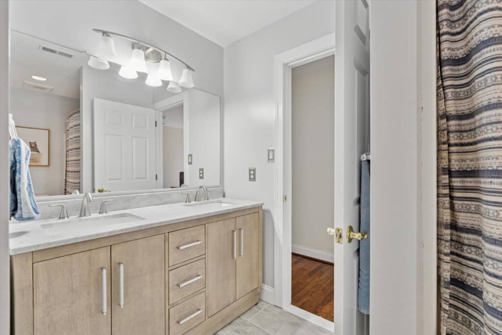Modern bathroom with a double-sink vanity, large mirror, light fixture above, and neutral-colored cabinets. A door is open to an adjacent room, and a striped shower curtain is partially visible on the right.
