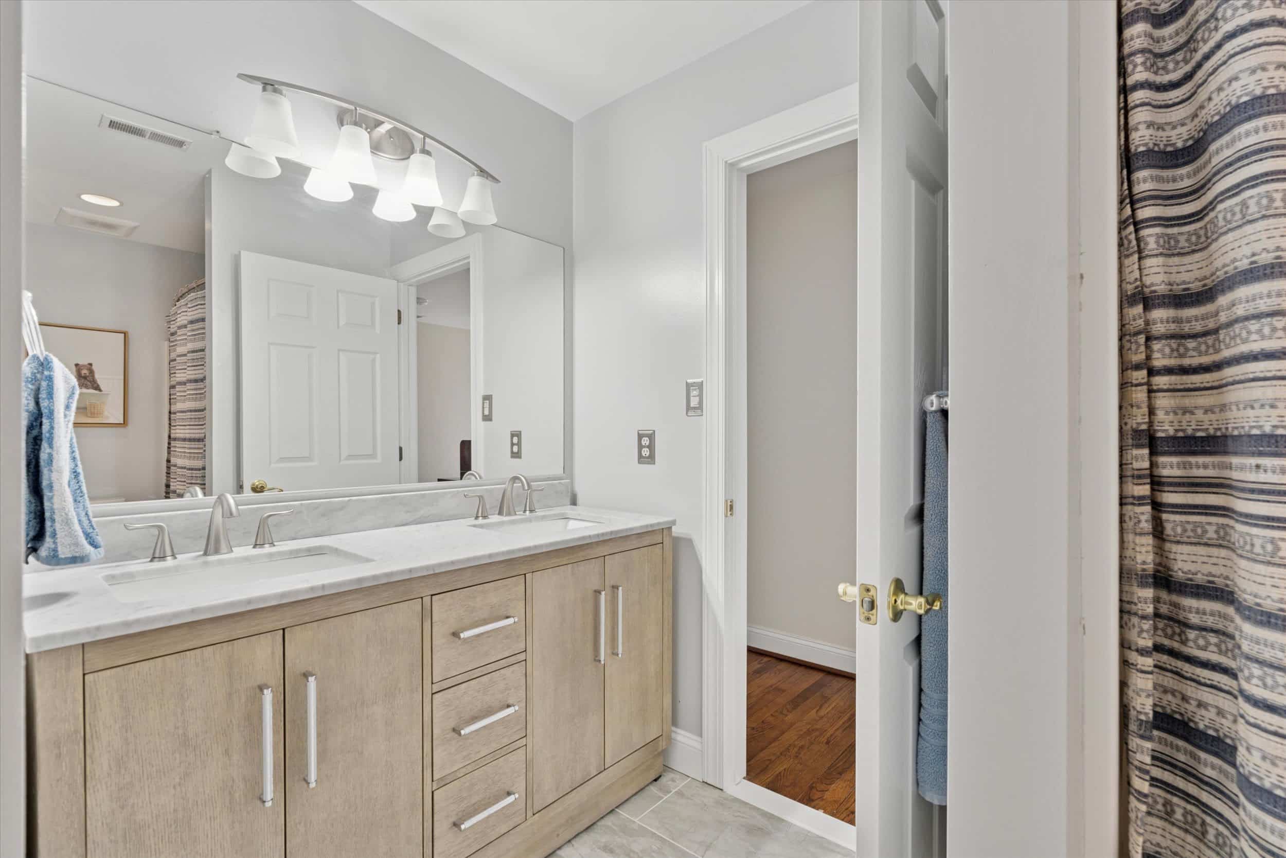Modern bathroom with a double-sink vanity, large mirror, light fixture above, and neutral-colored cabinets. A door is open to an adjacent room, and a striped shower curtain is partially visible on the right.