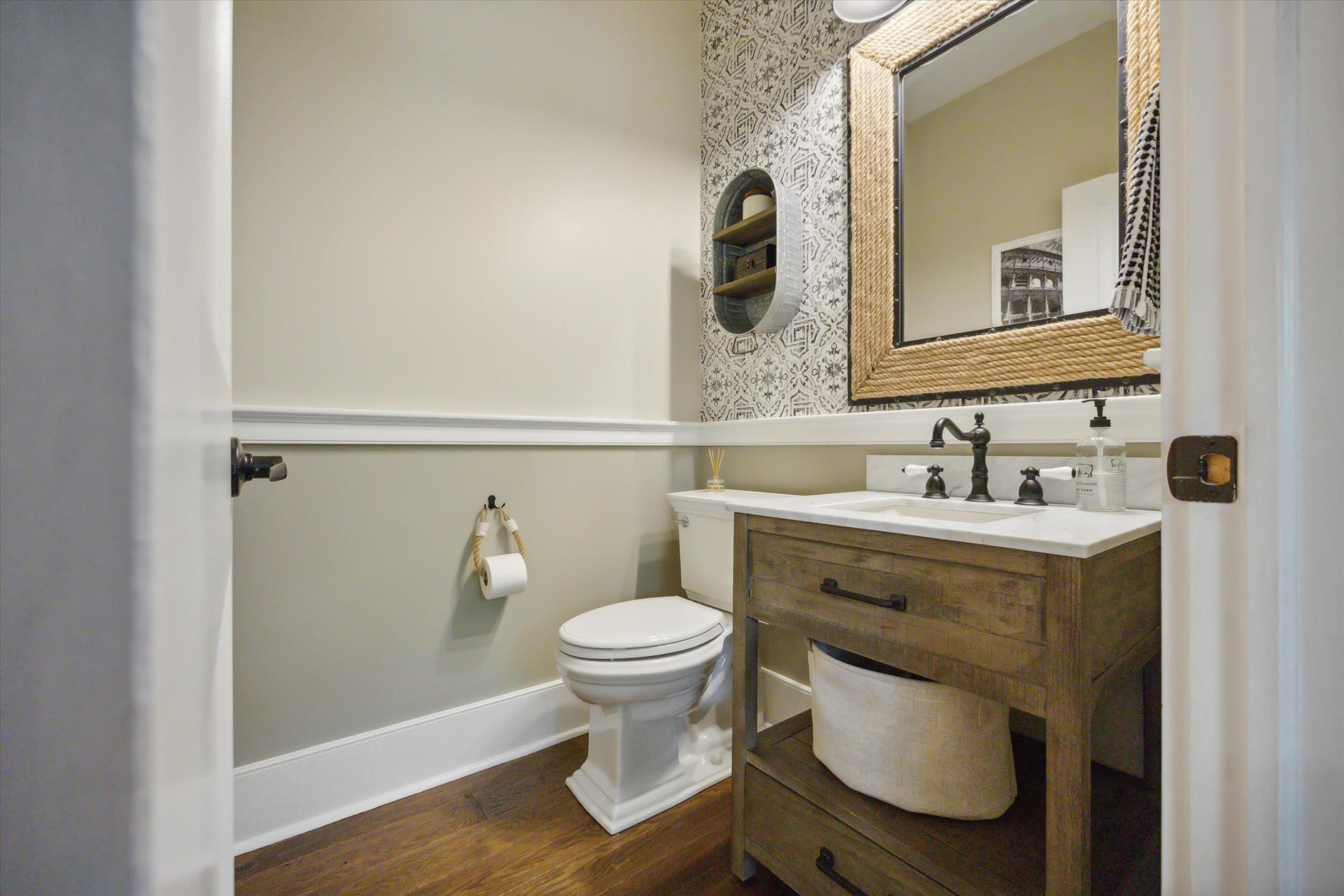 Bathroom-5 A small, modern bathroom with a wooden floor, a toilet, a wooden vanity with a sink, a large framed mirror, patterned wallpaper, and minimal decorative items on shelves and the vanity.