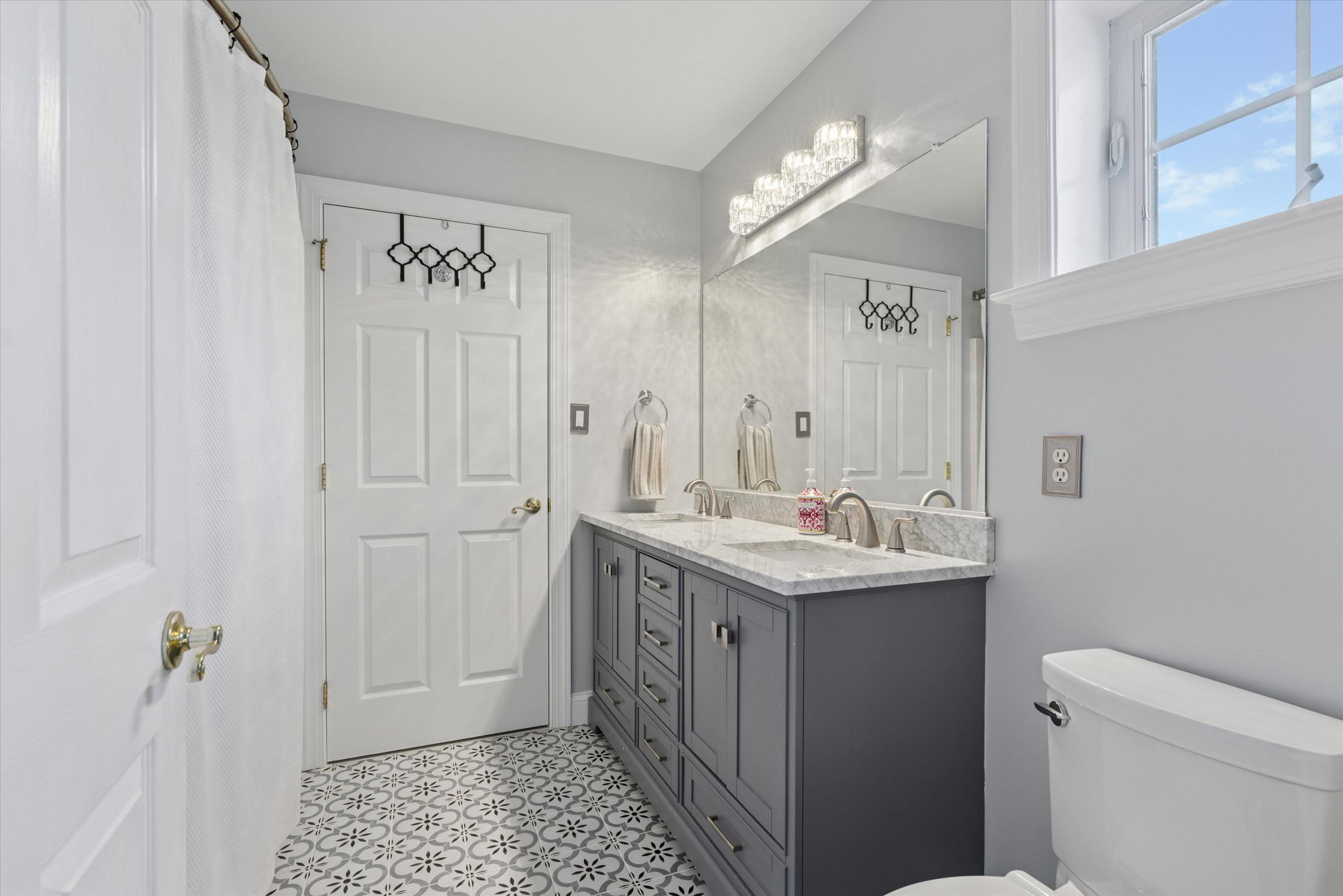 Modern bathroom with patterned tile floor, gray vanity with double sinks, marble countertop, large mirror, and bright lighting. White doors and a window let in natural light. A toilet is visible in the foreground.