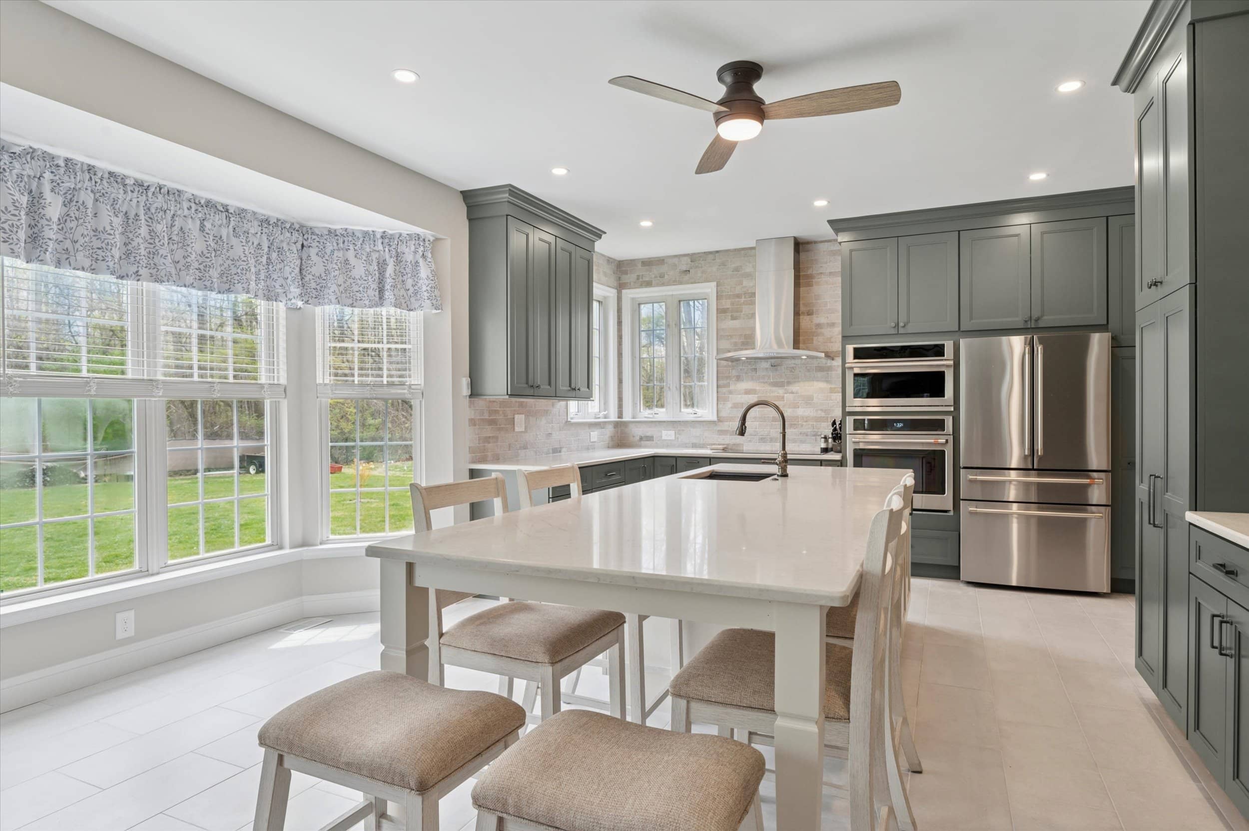 Photo-10 Bright, modern kitchen with sage green cabinets, stainless steel appliances, a white island with four upholstered stools, large bay windows with blue patterned curtains, and a ceiling fan.