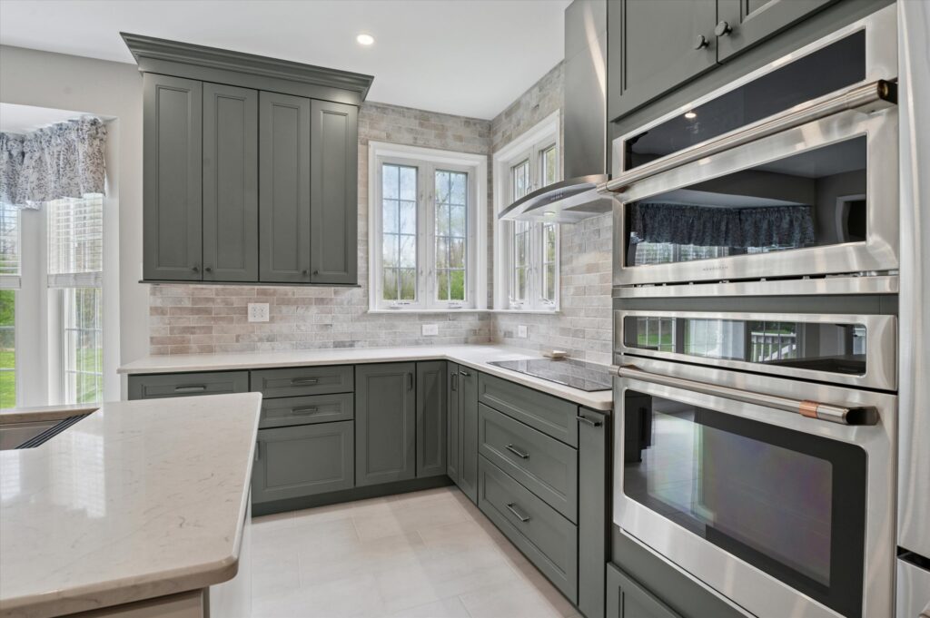 Photo-12 Modern kitchen with gray-green cabinets, marble countertops, stainless steel double ovens, and large windows allowing natural light to brighten the space. Light brick backsplash and neutral floor tiles complete the design.