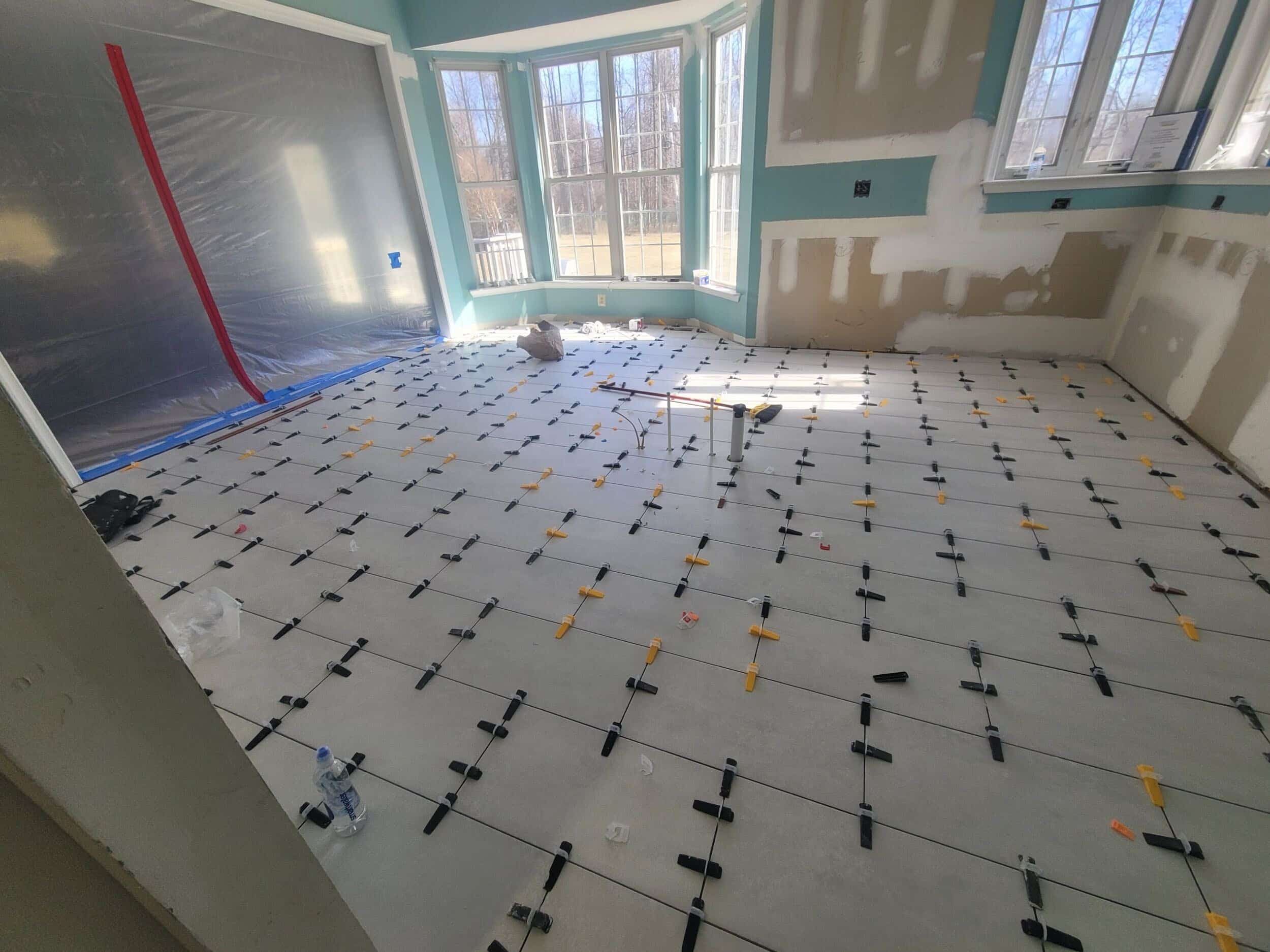Photo-5 A room with large windows is under renovation. Large square tiles are being installed on the floor, with spacers between them. The walls are partially unfinished, and construction tools are scattered around.