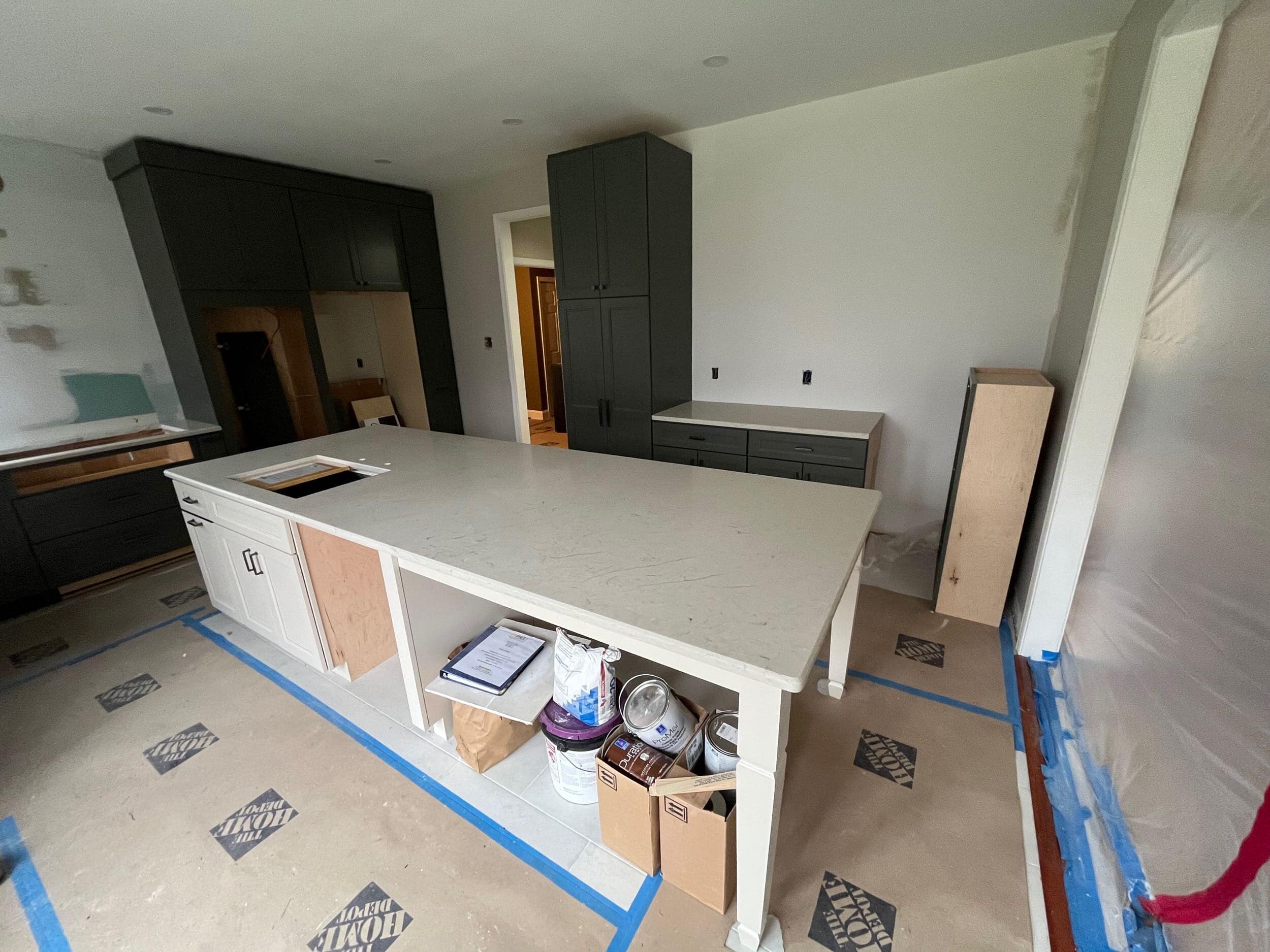 Photo-7 A modern kitchen under construction with a large island countertop, unfinished cabinets, and tools and supplies underneath the island. The floor is covered with protective material and blue painter’s tape lines the edges.