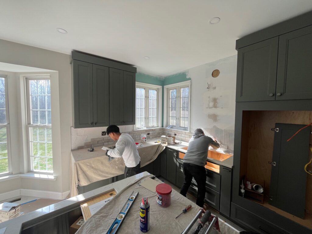 Photo-8 Two people are working on a kitchen renovation, painting and sanding near cabinets and a window. Tools, paint cans, and drop cloths are spread across the counters, and sunlight streams in through large windows.