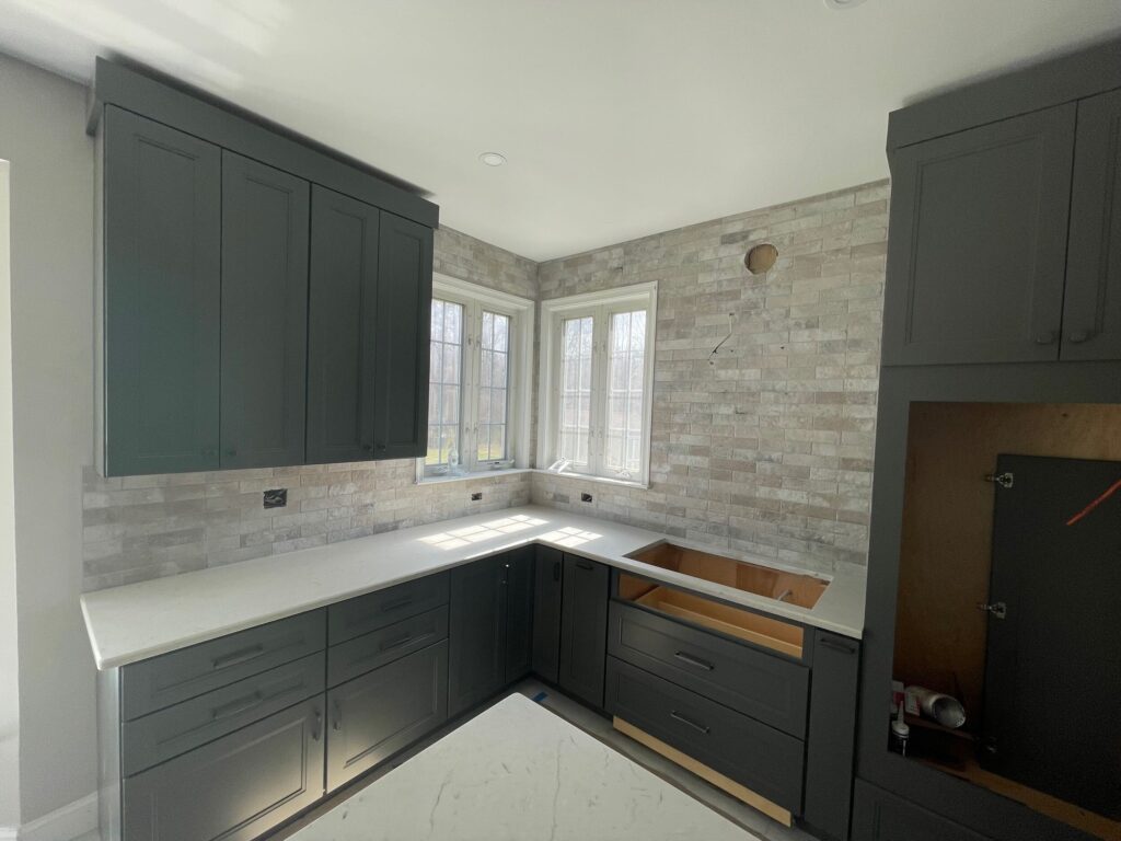 Photo-9 A kitchen corner with dark green cabinets, white countertops, and a light brick backsplash. There is a cabinet space left open, likely for an appliance, and sunlight is streaming through windows above the counter.