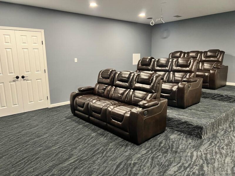 Home theater room with three rows of dark brown leather reclining seats on tiered carpeted flooring, gray-blue walls, a closed double door, and a ceiling-mounted projector.