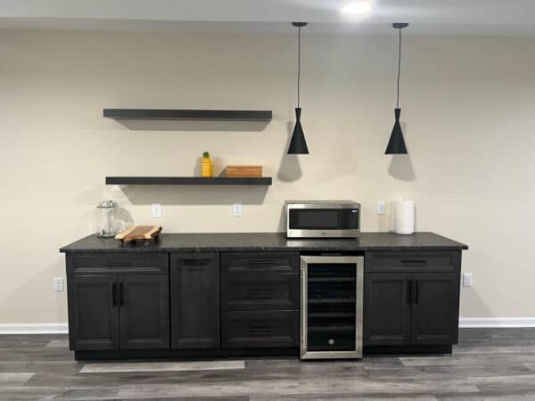 A modern kitchenette with dark cabinets, a black countertop, a microwave, a glass jar, a wine fridge, a paper towel roll, and two floating shelves holding a yellow bottle and a wooden box. Two black pendant lights hang above.